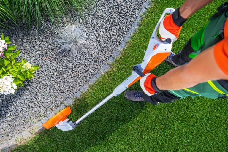 A person uses a string trimmer to neatly cut grass edges along a path in a well-maintained garden.